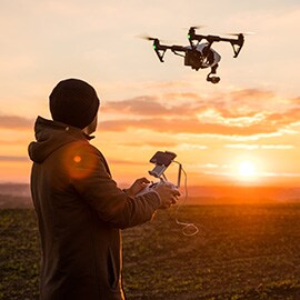A man outside using a two-handed remote to control a drone with a camera in the sky.