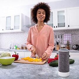 a woman chops vegetables in a kitchen while speaking a voice command to a smart speaker that is in the foreground of the photo.