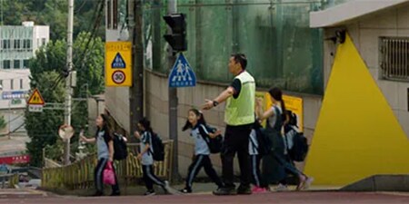 在群山環繞的城市裡，孩子們在身穿螢光安全背心的成年人的指揮下過馬路。
