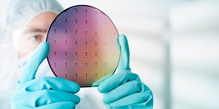 Photo of person in clean room holding a silicon wafer in gloved hands.

