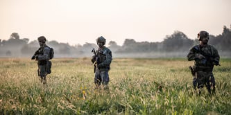 照片顯示，在晴朗的日子裡，六名身著全套戰術裝備的士兵站在一架軍用直升機附近的高草叢中。  
