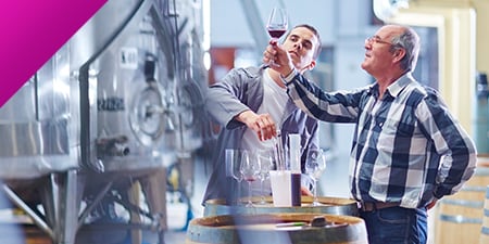 Two men inspecting a glass of wine in a winery.
