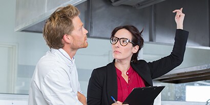 商業廚房裡，一名手持夾板的女性向上示意，廚師正注視著她。
