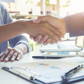 兩個人在桌上握手，桌上放著筆、文件夾和筆記型電腦。
