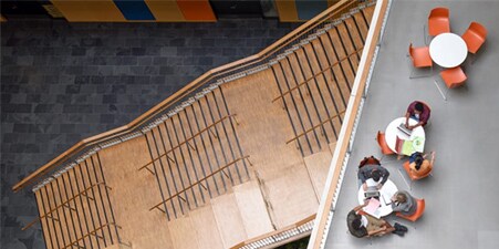 Bird's eye view or overhead shot of a school lobby with wood stairs and a seating section with orange chairs.
