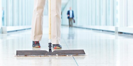 Cleaning person pushing a flat mop to clean debris from the floor of a school hallway.
