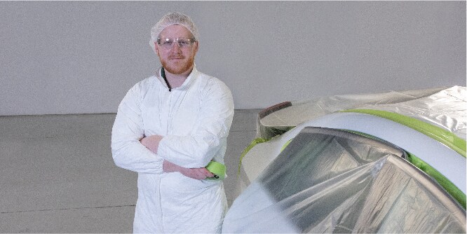 畫面為一位身穿白色工作服的工作人員，站在一輛用膠帶和遮蔽膠膜覆蓋的車輛旁邊。
