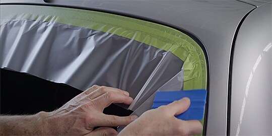 Video still with text “Wipe tape down with 3M™ Plastic Applicator PA-1” as a worker does that for tape on a window edge
