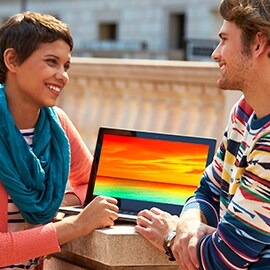 Two smiling people standing outside in front of a notebook computer that has a bright and colorful display screen.