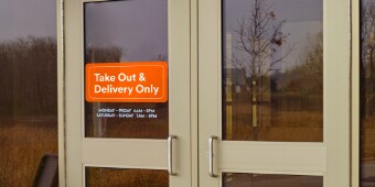 Orange window graphic on a restaurant’s front doors reading “Take Out & Delivery Only.”
