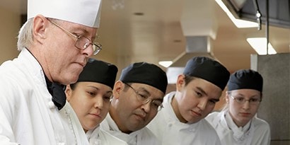 一位主廚正在示範食材準備技巧，四位廚房員工在旁觀看。
