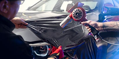 A graphic installer pulls Gloss Black 3M™ Wrap Film Series 2080 over a car hood while another installer heats it.

