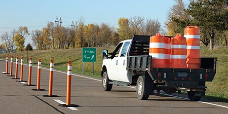 A truck carries orange and white plastic safety drums for highway construction.
