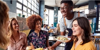 一名笑容可掬的服務生正在為餐廳內的四位用餐者送餐。
